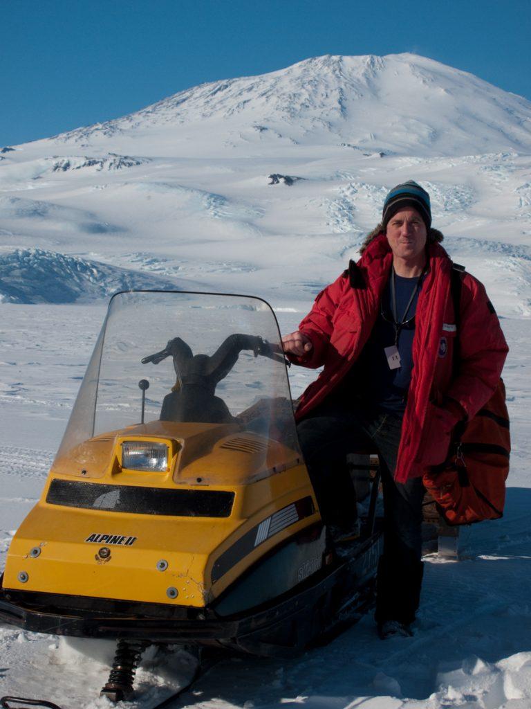 Doug Anderson stands next to a very old snow machine - Mount Erebus background. BBC Netflix Apple TV Wildlife Filming Filmmaking Underwater Natural History Ocean Cinematography Gates  Housings Nauticam REvo Rebreathers SCUBA Finistare clothing